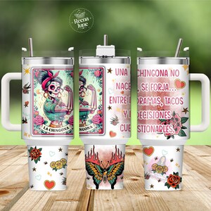 May include: A stainless steel tumbler with a handle and straw, featuring a graphic of a sugar skull woman in a tarot card style, a butterfly with flames, and the Spanish text: UNA CHINGONA NO NACE, SE FORJA... ENTRE PANORAMAS, TACOS Y DECISIONES CUESTIONABLES.