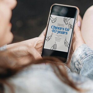 May include: A smartphone displays a birthday invitation with the text "Cheers to 40 years" in blue. The invitation includes line drawings of hands holding bottles and mugs. The phone is held by a person wearing a denim jacket.