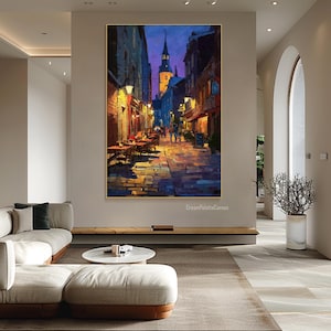 May include: An oil painting depicts a European street scene at dusk. The artwork features a cobblestone street, buildings with warm lighting, and a church steeple against a deep blue sky. Tables and chairs are set up outside restaurants.