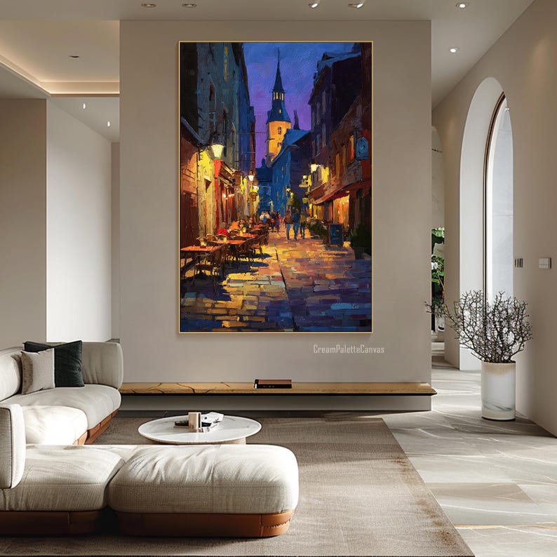 May include: An oil painting depicts a European street scene at dusk. The artwork features a cobblestone street, buildings with warm lighting, and a church steeple against a deep blue sky. Tables and chairs are set up outside restaurants.
