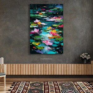 May include: An abstract painting of a pond with lily pads and flowers. The artwork features a vibrant color palette of pink, yellow, and green against a dark background. The painting is displayed on a gray wall above a wooden cabinet.