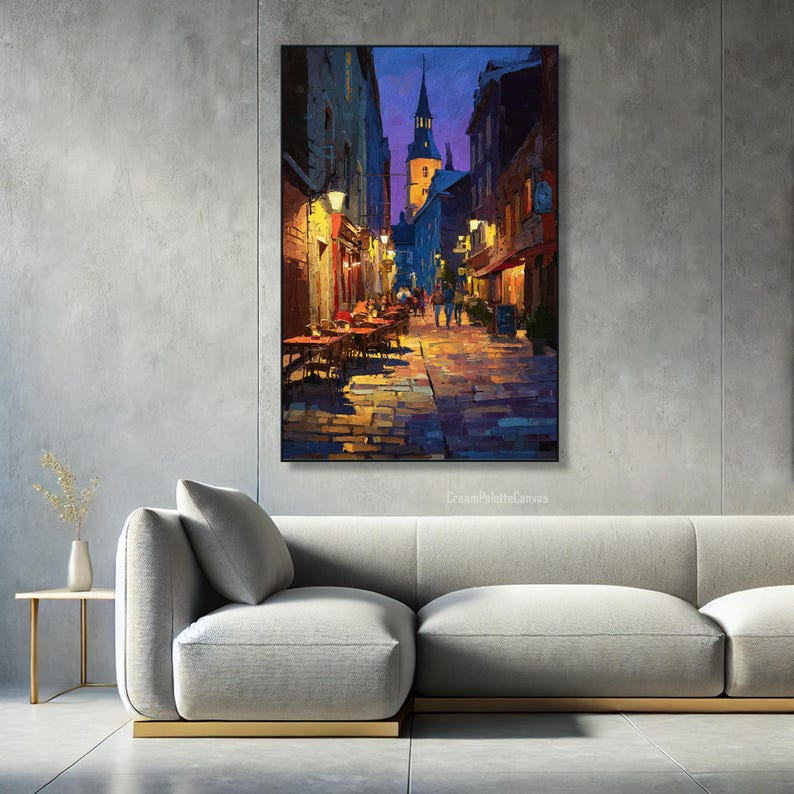 May include: An oil painting depicts a European street scene at dusk. The artwork features a cobblestone street illuminated by warm, inviting light from shops and street lamps. A tall steeple rises in the background, and cafe tables line the street.