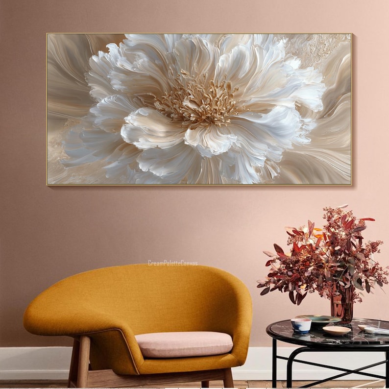 May include: A large, framed canvas art piece featuring a close-up of a white and beige flower with textured petals. The artwork has a gold frame and is displayed above a yellow armchair and a small table with a floral arrangement.