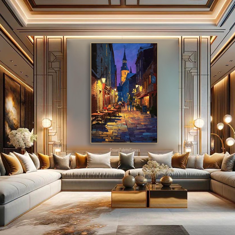 May include: An oil painting depicts a European street scene at dusk. The artwork features a cobblestone street, buildings with warm lighting, and a church steeple against a deep blue sky. The painting is displayed above a modern sofa.