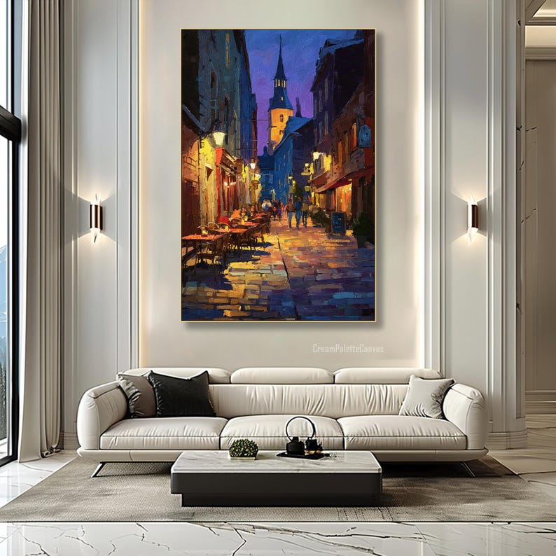 May include: An oil painting depicts a European street scene at dusk. The artwork features a cobblestone street, buildings with warm lighting, and a church steeple against a purple and blue sky. Tables and chairs are set up outside.