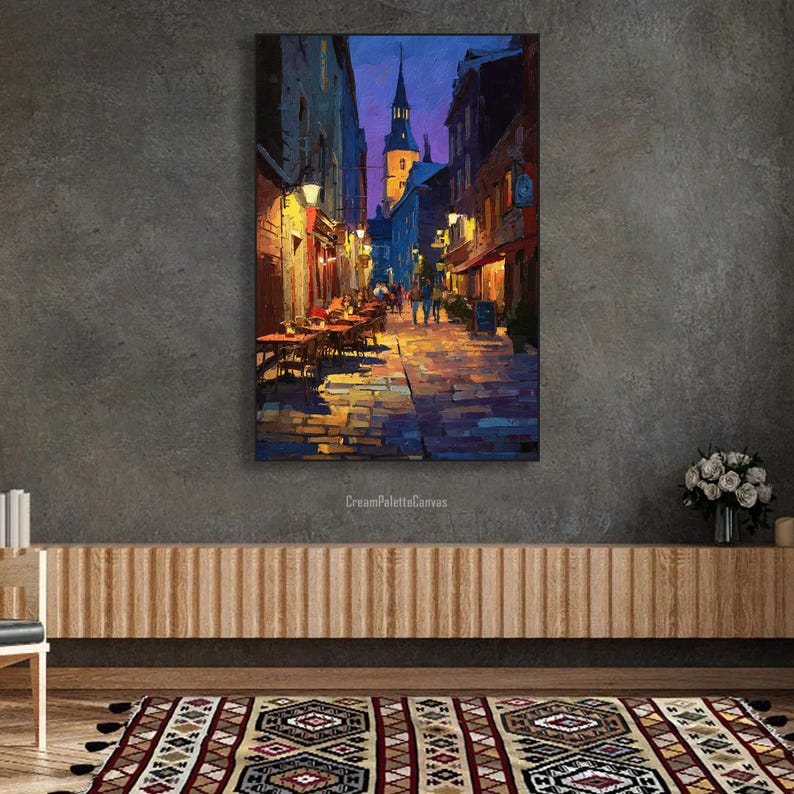 May include: An oil painting depicts a European street scene at dusk. The artwork features a cobblestone street lined with buildings, illuminated by warm streetlights. Tables and chairs are set up outside restaurants. A church steeple rises in the distance.