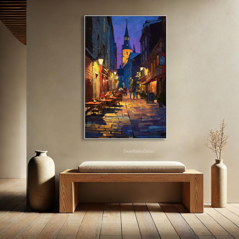 May include: An impressionistic painting of a European street scene at dusk. The artwork depicts a cobblestone street lined with buildings, restaurants, and a church tower. Warm light from street lamps and shop windows contrasts with the cool blues and purples of the evening sky.