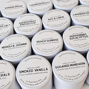 May include: A collection of 16 white candle tins with black labels and various scents. The labels include names like "Aloe & Bamboo", "Banana Bread", "Caribbean Nights", "Coconut Lime", "Fig Grove", "Freesia & Sea Salt", "Grapefruit Prosecco", "Iris & Linen", "Lavender Dreams", "Lemon Verbena", "Neroli & Jasmine", "Peach Sunrise", "Peppermint Eucalyptus", "Rosemary Mint", "Sea Minerals", "Smoked Vanilla", "Sugar Mandarine", "Sunkissed Cotton".