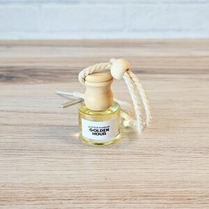 Puede incluir: Una peque&ntilde;a botella de vidrio con una tapa de madera y una etiqueta blanca que dice "good faith handmade GOLDEN HOUR". La botella est&aacute; colgada de una cuerda blanca con una cuenta de madera.