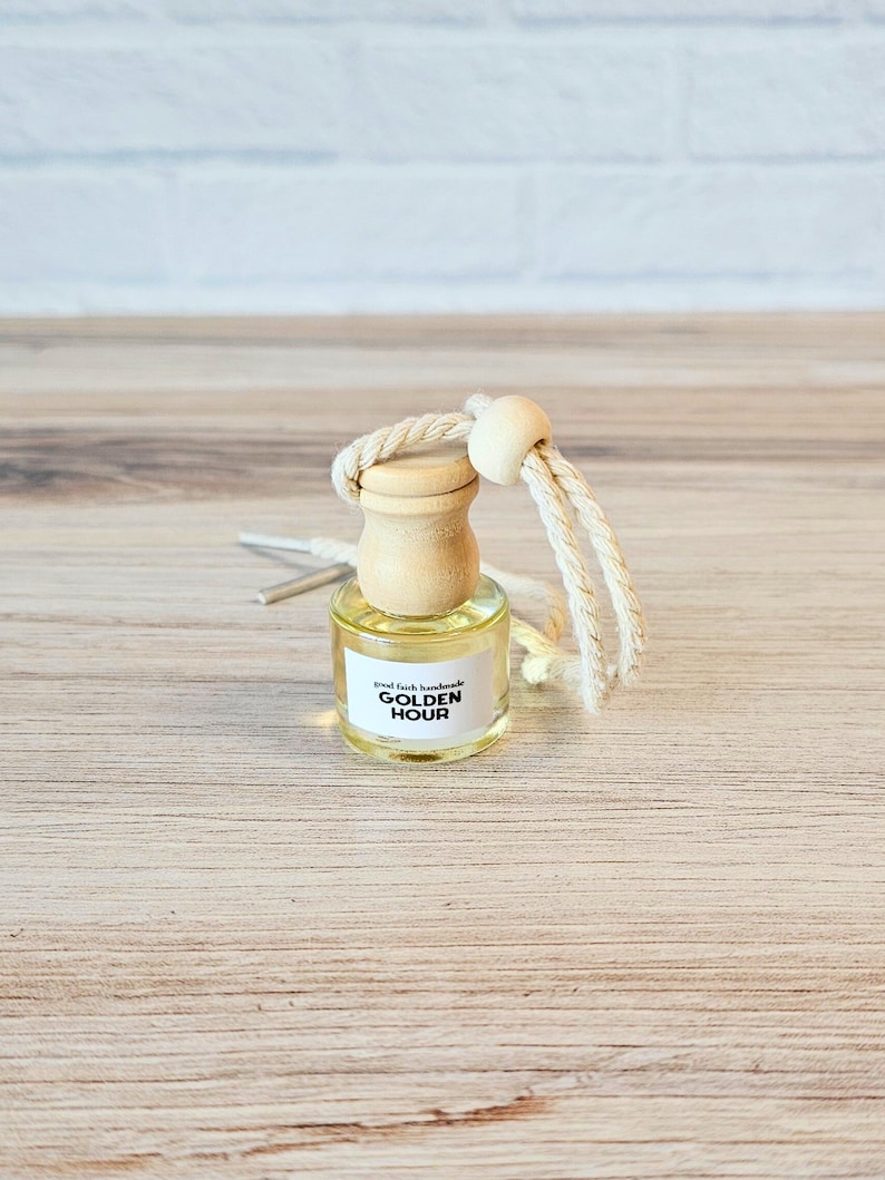 Puede incluir: Una peque&ntilde;a botella de vidrio con una tapa de madera y una etiqueta blanca que dice "good faith handmade GOLDEN HOUR". La botella est&aacute; colgada de una cuerda blanca con una cuenta de madera.