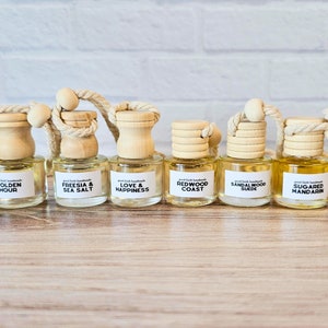 May include: Ten small glass bottles with wooden caps and string hangers. Each bottle contains a different scented oil and has a label with the scent name. The scents are Coconut Lime, Golden Hour, Freesia & Sea Salt, Love & Happiness, Redwood Coast, Sandalwood Suede, Sugared Mandarin, and Sun Kissed Cotton.