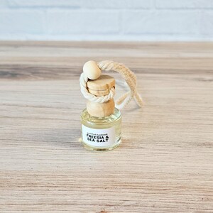 Puede incluir: Una peque&ntilde;a botella de vidrio con una tapa de madera y una cuerda para colgar. La botella est&aacute; llena de un l&iacute;quido transparente y tiene una etiqueta que dice "Good Faith Handmade Freesia & Sea Salt".