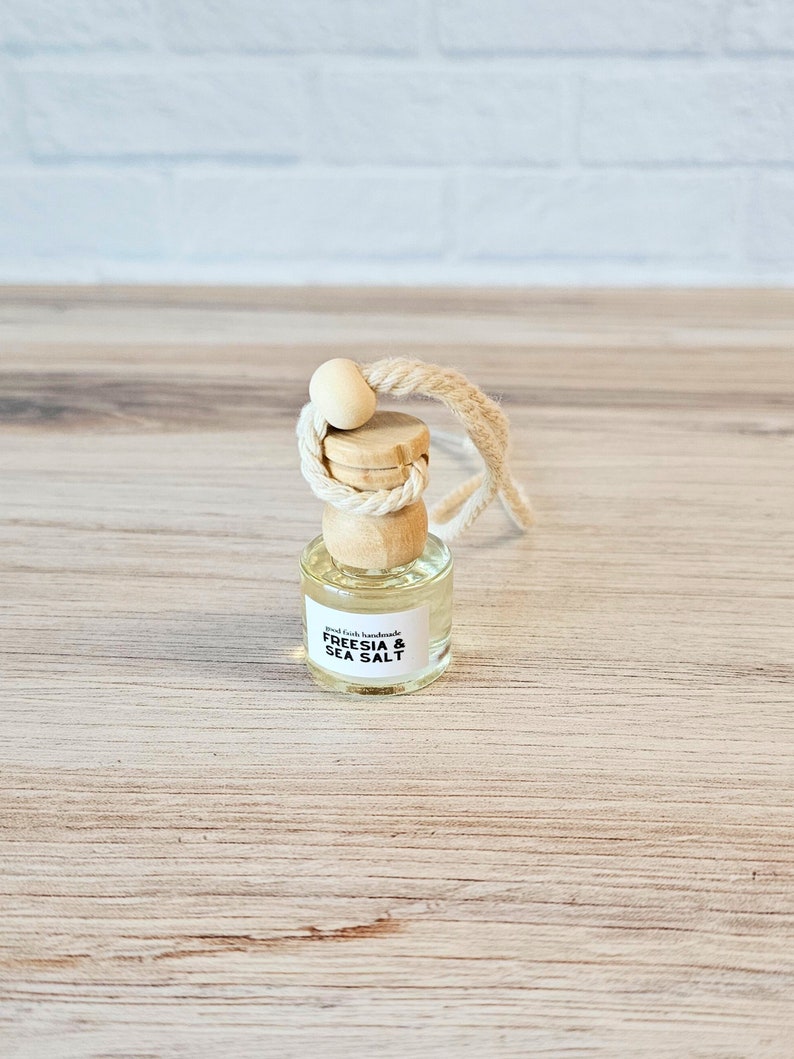 Puede incluir: Una peque&ntilde;a botella de vidrio con una tapa de madera y una cuerda para colgar. La botella est&aacute; llena de un l&iacute;quido transparente y tiene una etiqueta que dice "Good Faith Handmade Freesia & Sea Salt".