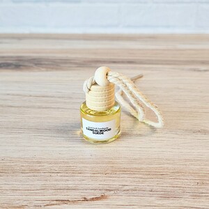 Puede incluir: Una peque&ntilde;a botella de vidrio con un tap&oacute;n de corcho y una etiqueta blanca que dice "Small Batch Handmade Sandalwood Suede". La botella est&aacute; colgada de una cuerda blanca.