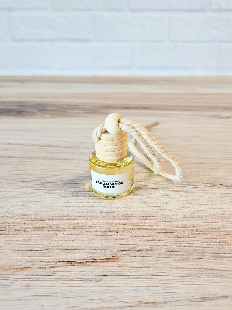 Puede incluir: Una peque&ntilde;a botella de vidrio con un tap&oacute;n de corcho y una etiqueta blanca que dice "Small Batch Handmade Sandalwood Suede". La botella est&aacute; colgada de una cuerda blanca.