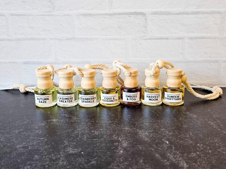 Clear glass car diffuser with wooden lid designed to hang from rearview mirror. Fall scents pictured are Autumn Daze, Cashmere Sweater, Cranberry Sparkle, Cider & Doughnuts, Embers & Fog, Harvest Moon, and Pumpkin Everything.