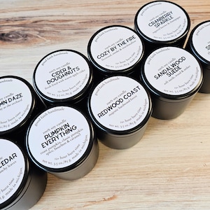 May include: Ten black candle jars with white labels. Each label has a different scent name and description. The scents include Autumn Daze, Leaves & Cedar, Pumpkin Everything, Cider & Doughnuts, Cozy by the Fire, Cranberry Sparkle, Redwood Coast, Sandalwood Suede, Spiced Plum.