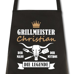 Könnte beinhalten: Schwarze Schürze mit dem Text "GRILLMEISTER Christian" in Weiß und Gold. Darunter ein Bullenschädel mit gekreuzten Grillwerkzeugen und den Worten "DER MANN DER MYTHOS DIE LEGENDE". Oben eine goldene Krone.