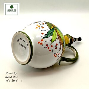May include: White ceramic olive oil bottle with a green handle and a yellow spout. The bottle is decorated with hand-painted green leaves, orange dots, and black swirls. The bottom of the bottle reads "Fatto a Mano Firenze".