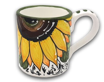 May include: A white ceramic mug with a green rim and a sunflower design. The sunflower is painted in shades of yellow, orange, brown, and green. The mug has a white handle with black polka dots.