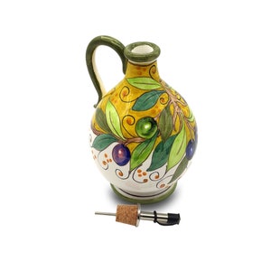 May include: A white ceramic olive oil bottle with a green handle and a yellow, green, and blue olive branch design. The bottle has a cork stopper and a metal spout.