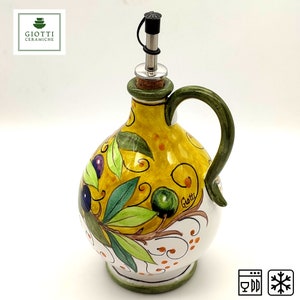 May include: Yellow ceramic olive oil bottle with a green handle and a silver spout. The bottle is decorated with green leaves, brown branches, and purple and green olives. The bottle has the text "Giotti" on the side.