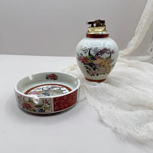 Pode incluir: Um conjunto vintage de cinzeiro e isqueiro em porcelana. O cinzeiro é redondo com uma borda vermelha e um desenho central de pavão. O isqueiro é branco com um topo dourado e um desenho de pavão a condizer. O conjunto é provavelmente da meados do século XX.