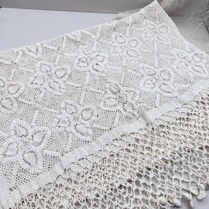 Può includere: Tessuto in pizzo color crema fatto a mano con un motivo floreale. Il pizzo presenta un design dettagliato con un bordo a frange. Il tessuto è piegato, mostrando l'intricata maestria e la consistenza del tessuto delicato.