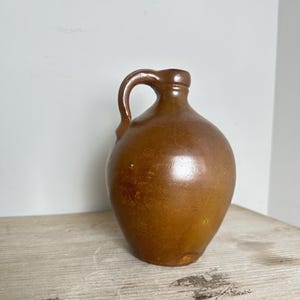 May include: A brown ceramic jug with a rounded body, a narrow neck, and a single curved handle attached to the side. The surface has a smooth, glazed finish with subtle variations in color and small imperfections visible on the exterior.