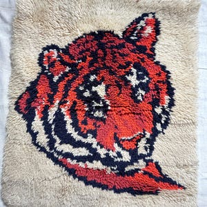 May include: A beige, rectangular rug featuring a tiger's head design. The tiger's face is rendered in shades of red, black, and white, with a shaggy texture. The rug's background is a neutral beige color.