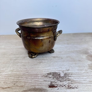 Puede incluir: Un pequeño cuenco metálico redondo con acabado en latón desgastado, que cuenta con dos asas laterales y tres patas cortas decorativas. El recipiente tiene una abertura ancha y un cuerpo curvo con signos visibles de oxidación y color.