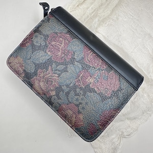 May include: A black and floral wallet with a leather texture. The wallet features a pattern of pink and purple roses with blue-green leaves. The top of the wallet is black.