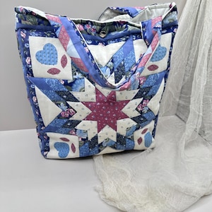 Peut inclure: Sac fourre-tout matelassé avec un motif patchwork. Le sac présente une étoile centrale rose et blanche, entourée de carrés bleus et blancs avec des motifs de cœurs et de fleurs. Les poignées et la bordure sont bleu uni.