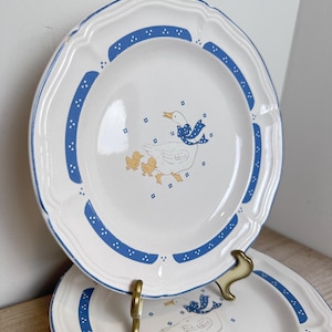 May include: White ceramic plates with a blue and white scalloped edge design. Each plate features a blue band with white dots and a central illustration of a goose with a blue bow and yellow ducklings. Decorative tableware.