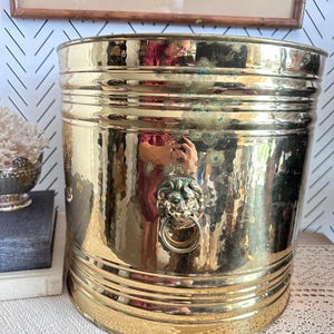 Könnte beinhalten: Ein großer, gehämmertes Messing-Eimer mit einem Löwenkopf-Ringgriff. Der Eimer hat eine zylindrische Form mit horizontalen Bändern und einer strukturierten Oberfläche. Das Messing hat einen warmen, goldenen Farbton mit Patina.