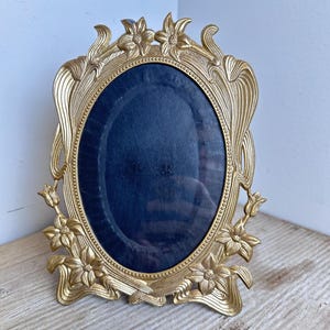 May include: An oval picture frame with a metallic gold finish, featuring ornate floral and leaf relief detailing along the edges. The central display area is covered by a dark navy blue fabric backing, surrounded by a beaded inner border design.