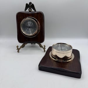 Puede incluir: Dos instrumentos meteorológicos antiguos de madera. Uno es un barómetro con un águila de latón, montado sobre un soporte. El otro es una brújula sobre una base de madera. Ambos tienen detalles de latón, sobre fondo blanco.