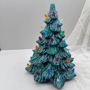 Puede incluir: Un árbol de Navidad de cerámica de color verde turquesa con luces de colores. El árbol tiene una superficie texturizada y está decorado con luces multicolores, incluyendo rojo, amarillo, azul y verde.