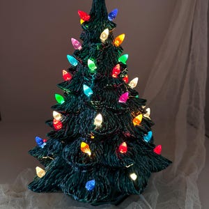 May include: A ceramic Christmas tree in a dark green glaze, decorated with multi-coloured lights. The lights are teardrop-shaped, in shades of red, yellow, blue, green, and pink. A white electrical cord emerges from the base.