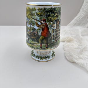 May include: A ceramic pedestal cup with a gold rim and base. The cup features a detailed illustration of a person hunting in a rural setting. The word "FRIENDSHIP" is written around the base.