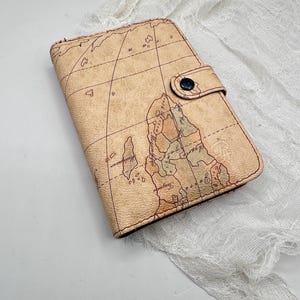 May include: A tan-colored wallet with a vintage map design. The wallet has a flap closure with a black button. The map design features continents, grid lines, and various geographical details. The wallet is closed and appears to be made of a leather-like material.