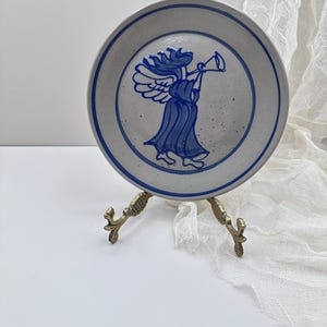 May include: A ceramic plate featuring a blue angel design. The plate has a light grey background, a blue border, and a blue illustration of an angel playing a trumpet. The plate is displayed on a decorative gold-coloured stand.