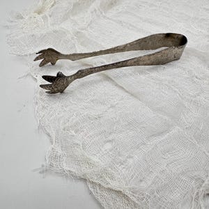 May include: Antique silver-toned sugar tongs with bird claw-shaped ends. The tongs are resting on a white, textured fabric. The tongs are designed to pick up sugar cubes or other small items.