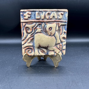 May include: A ceramic tile with a relief design of a cat. The tile is decorated with blue, red, and white glaze. The word "LYCAS" is written in red on the top of the tile.