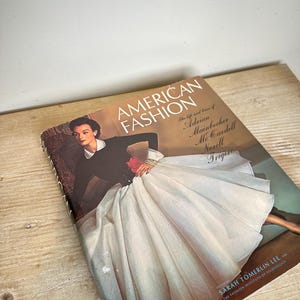 Può includere: Un libro con copertina rigida intitolato American Fashion, a cura di Sarah Tomerlin Lee. La copertina presenta una fotografia di una persona con un top nero a maniche lunghe e un'ampia gonna plissettata color bianco sporco, con il testo che elenca i designer Adrian, Mainbocher, McCardell, Norell e Trigere.