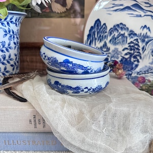Könnte beinhalten: Zwei kleine, gestapelte, weiß-blaue Keramikschalen mit einem Drachendesign. Die Schalen sind Teil einer Sammlung aus blau-weißem Porzellan, darunter eine Vase und ein dekorativer Teller. Die Schalen liegen auf einem Stapel Bücher und einem weißen Tuch.