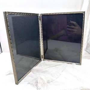 May include: An antique, hinged photo frame with a silver-colored metal frame and black backing. The frame is open, revealing two sections. The edges of the frame have a decorative, ornate design. The frame is set against a white background.