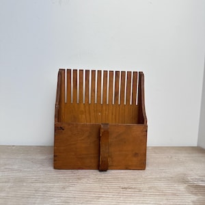 May include: A vintage wooden berry picker, featuring a handle and vertical slats. The rectangular box is crafted from light brown wood, with a series of vertical slats at the top. The handle is attached to the front.