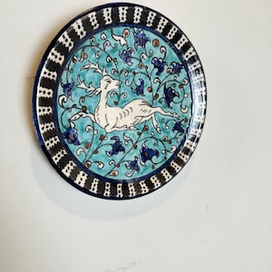 Op de afbeelding: Een ronde keramische schaal met een centrale witte hertenillustratie tegen een levendige turquoise achtergrond met donkerblauwe bloemmotieven. De schaal is omlijst door een brede rand met een herhalend zwart-wit verticaal streeppatroon.