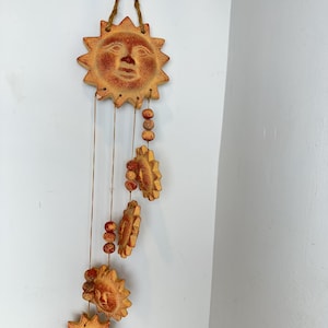 Könnte beinhalten: Eine hängende Wanddekoration mit fünf terrakottafarbenen Sonnengesichtern mit strukturierter Oberfläche. Die Sonnen sind mit dünner Schnur verbunden und mit kleinen runden Perlen durchsetzt, aufgehängt an einer Schlaufe aus Jutekordel.